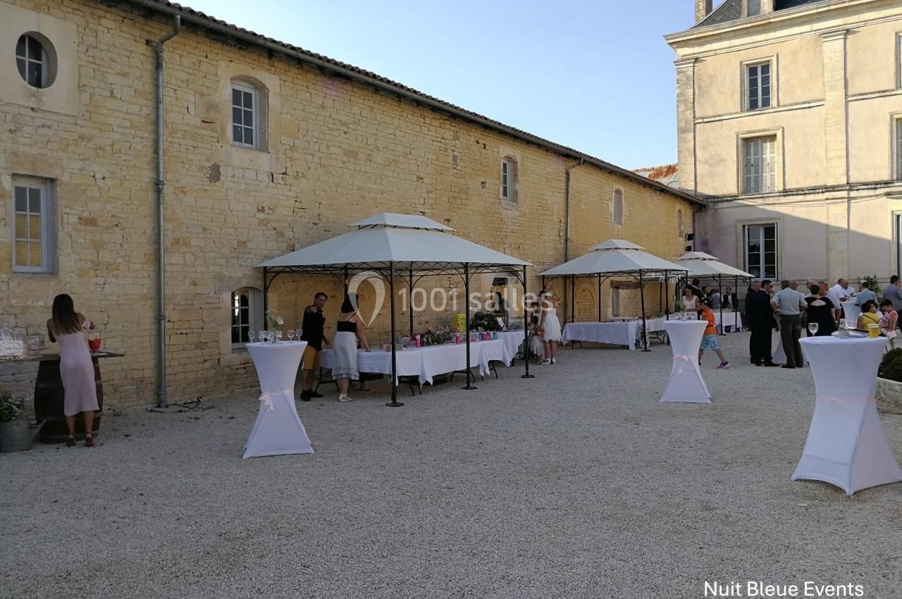 Cour extérieure avec des tables sous des tonnelles, des invités debout et un bâtiment en pierre en arrière-plan.