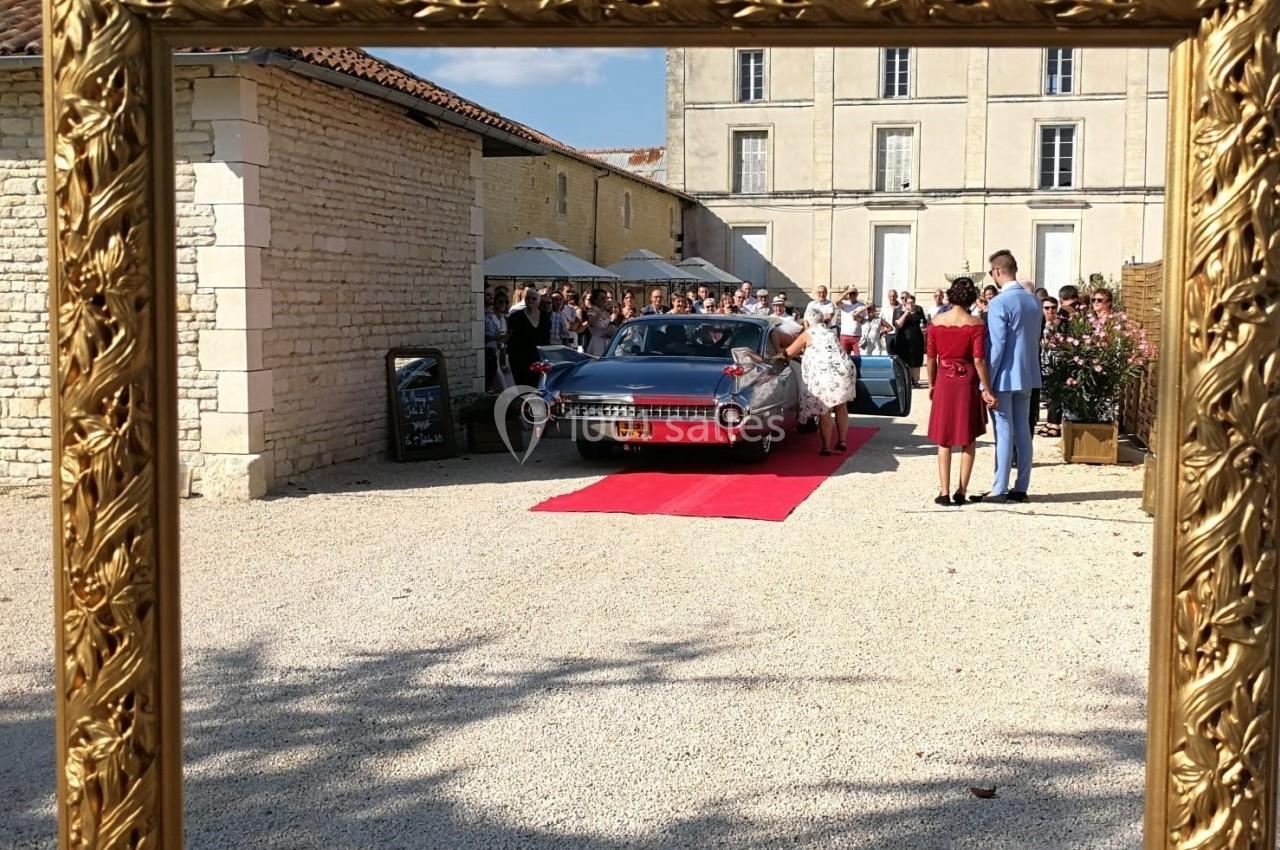 Cadre doré entourant une scène de mariage avec une voiture ancienne, un tapis rouge et des invités devant un bâtiment en…