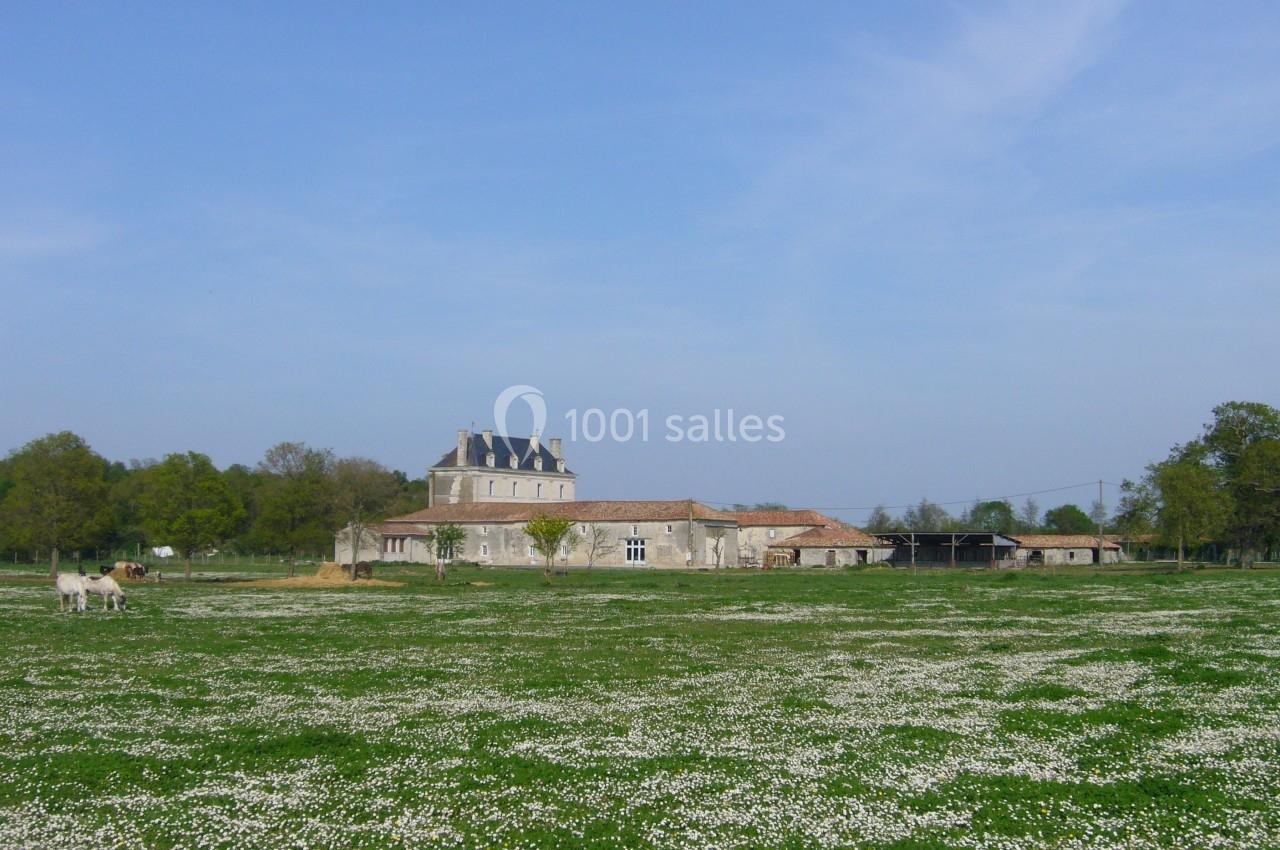 Bâtisse en pierre entourée de prairies fleuries sous un ciel bleu, avec des arbres en arrière-plan.