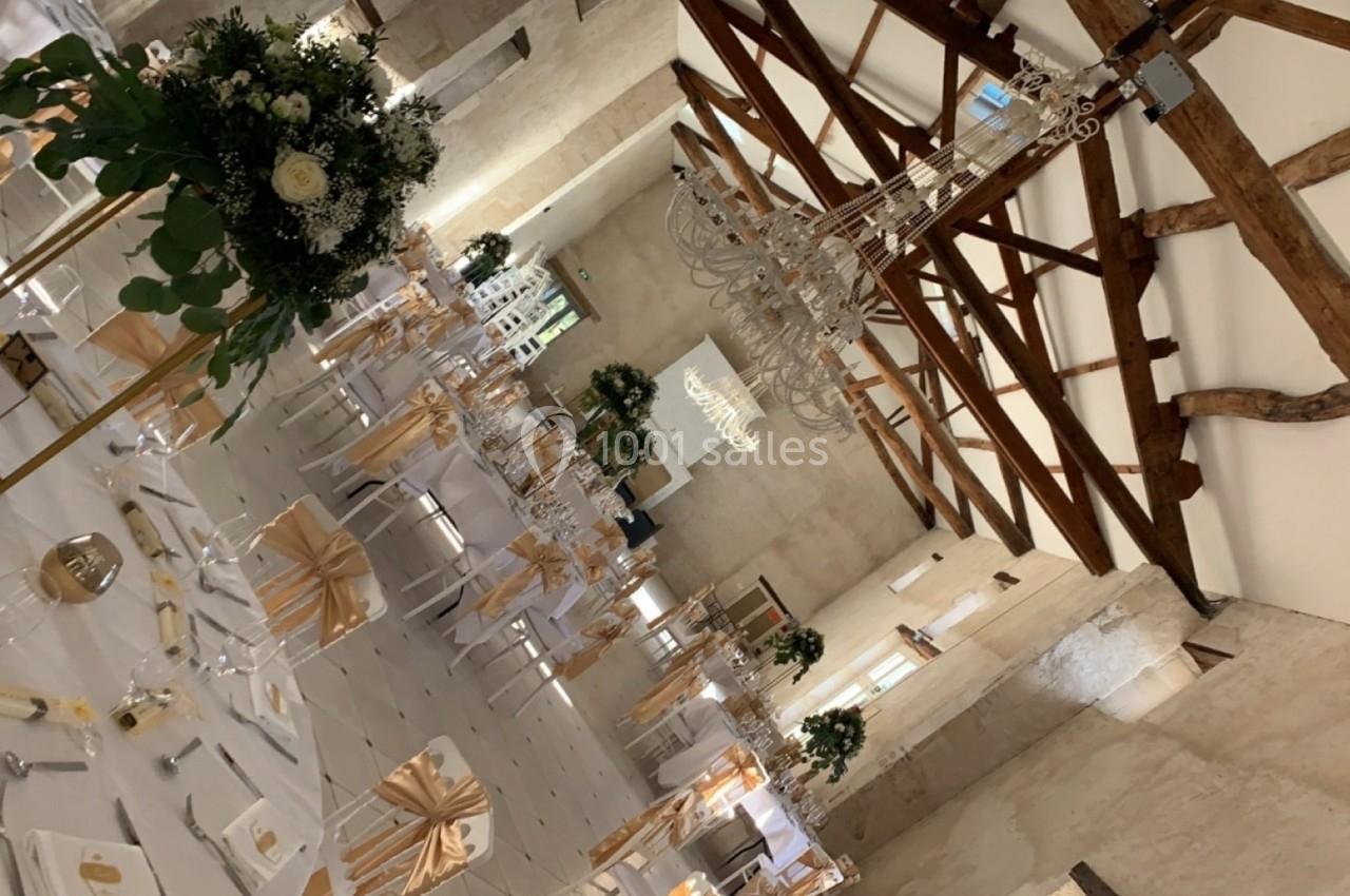 Salle de réception décorée pour un mariage, avec tables dressées, chaises ornées de nœuds dorés et lustres suspendus.