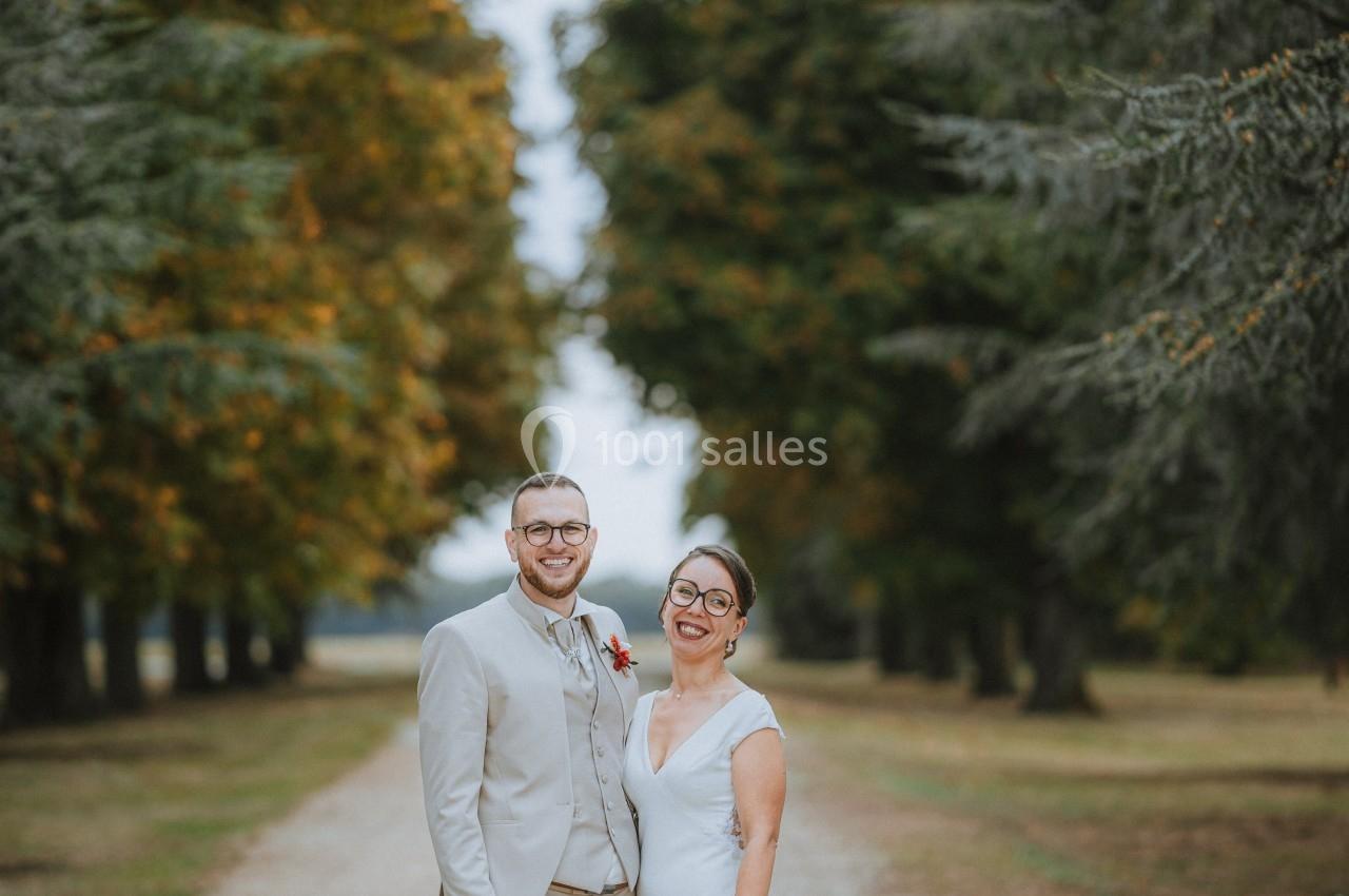 Un couple en tenue de mariage pose sur une allée bordée d'arbres dans un cadre naturel.