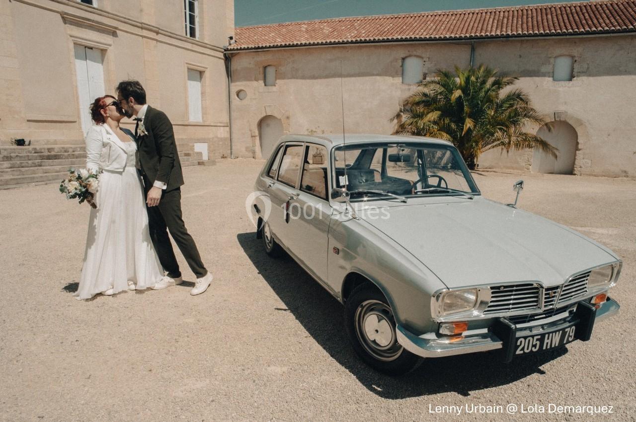 Un couple de mariés s'embrasse devant une voiture ancienne stationnée dans une cour ensoleillée.