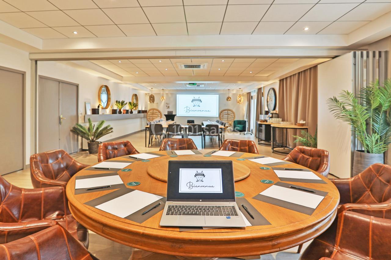 Salle de réunion lumineuse avec table ronde, chaises en cuir, ordinateur portable et écran de présentation.