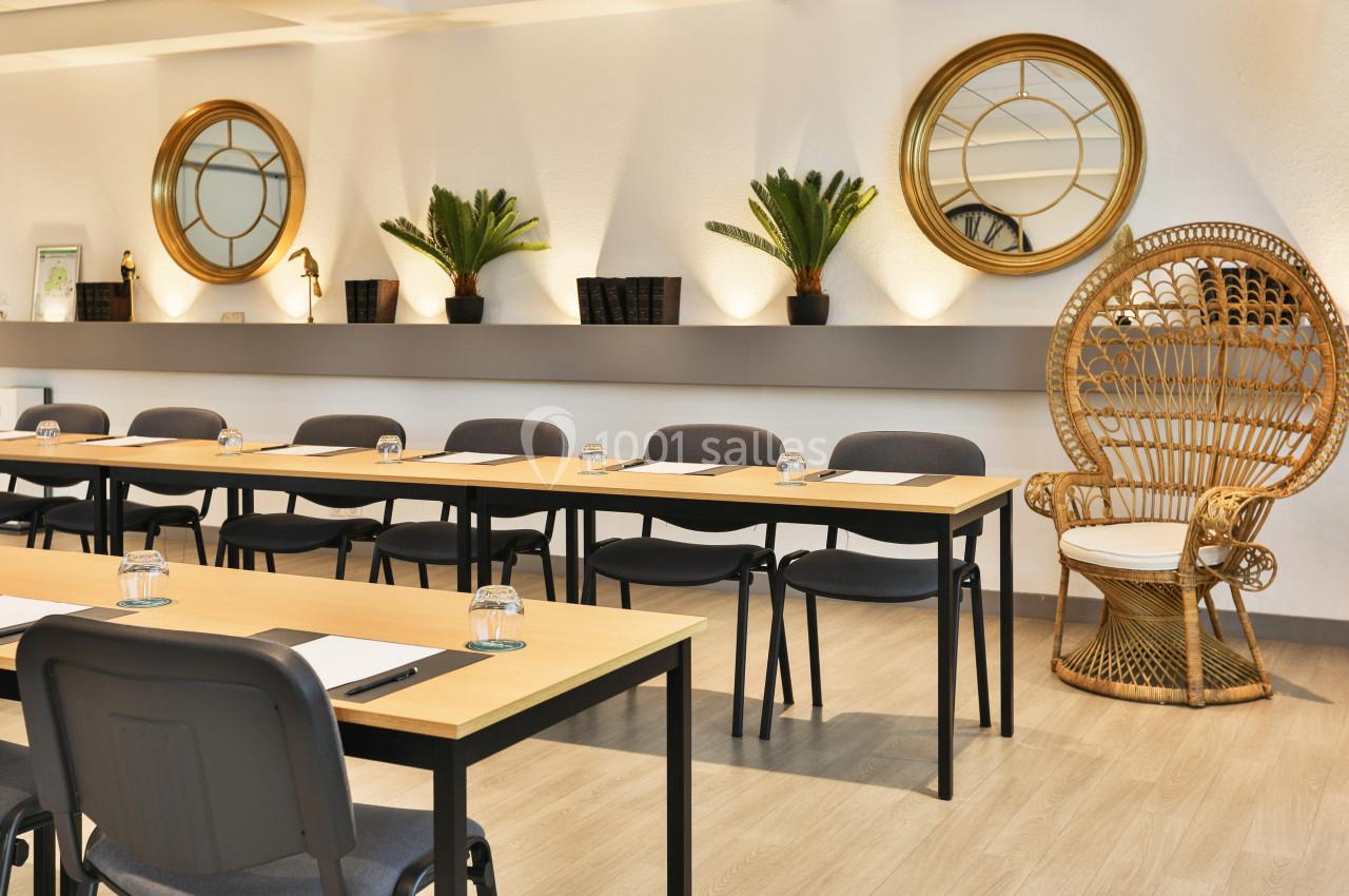 Salle de réunion lumineuse avec tables en bois, chaises noires, miroirs ronds, plantes et fauteuil en rotin.