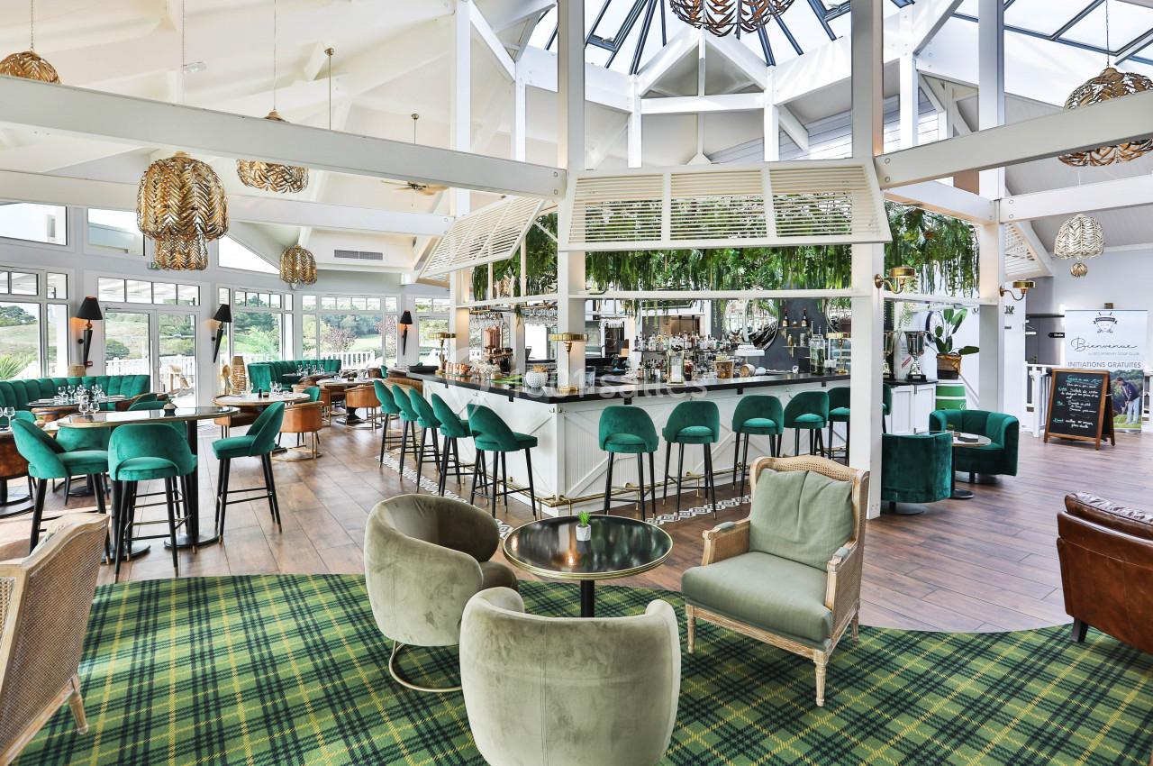 Intérieur lumineux d'un bar-restaurant avec mobilier vert, comptoir central, plantes suspendues et grandes baies vitrées.