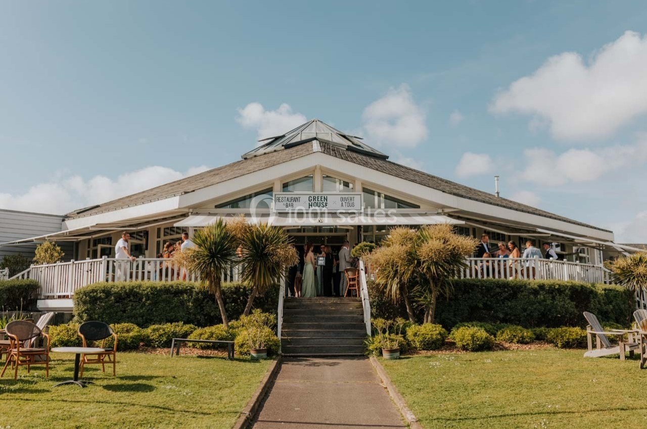 Location salle Talmont-Saint-Hilaire (Vendée) - Bourgenay Golf Club #1 Bâtiment lumineux avec terrasse en bois, entouré de palmiers et de verdure, sous un ciel bleu dégagé.