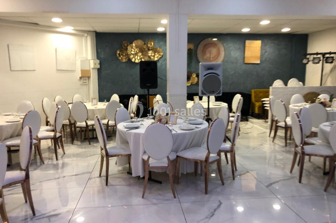 Salle de réception avec tables rondes dressées, chaises blanches et décoration murale dorée sur fond bleu.