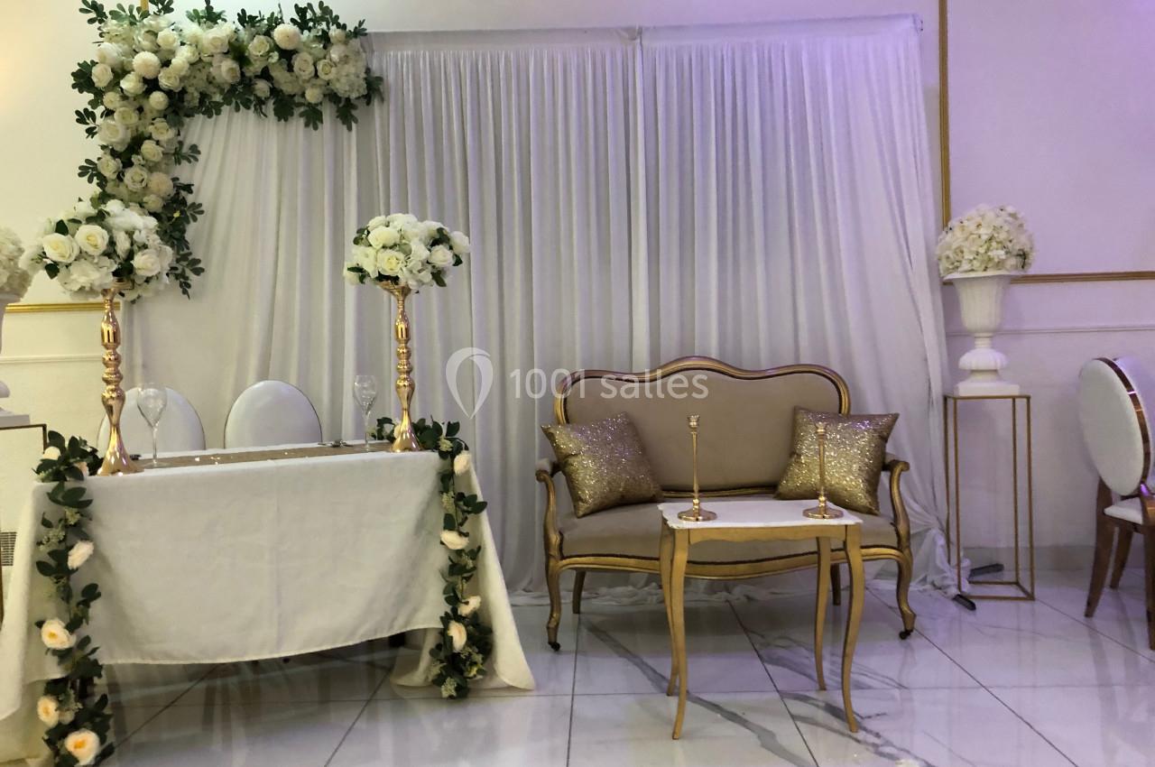 Décoration de salle avec table ornée de fleurs blanches, rideau blanc en fond et canapé doré avec coussins scintillants.