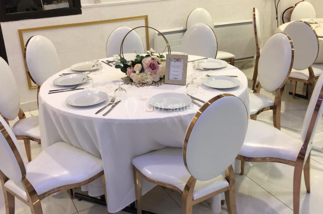 Table ronde élégamment dressée avec nappes blanches, assiettes, couverts et centre de table floral dans une salle lumineuse.