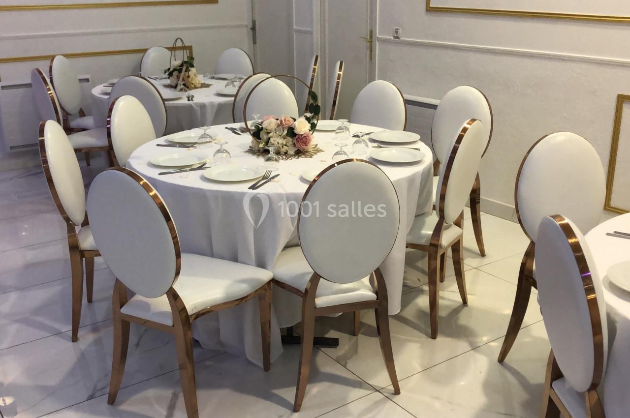 Salle décorée avec des tables rondes dressées, nappes blanches, chaises élégantes et centre de table floral.