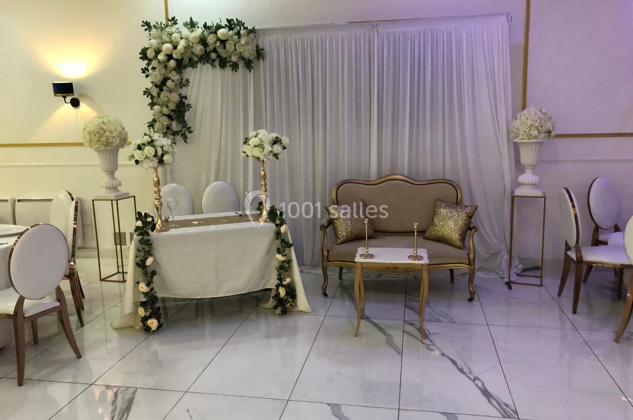 Salle de réception décorée avec des fleurs blanches, une table ornée et un canapé doré devant un rideau blanc.