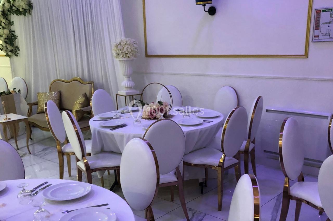 Salle de réception élégante avec tables dressées, chaises blanches et décorations florales dans un cadre lumineux.