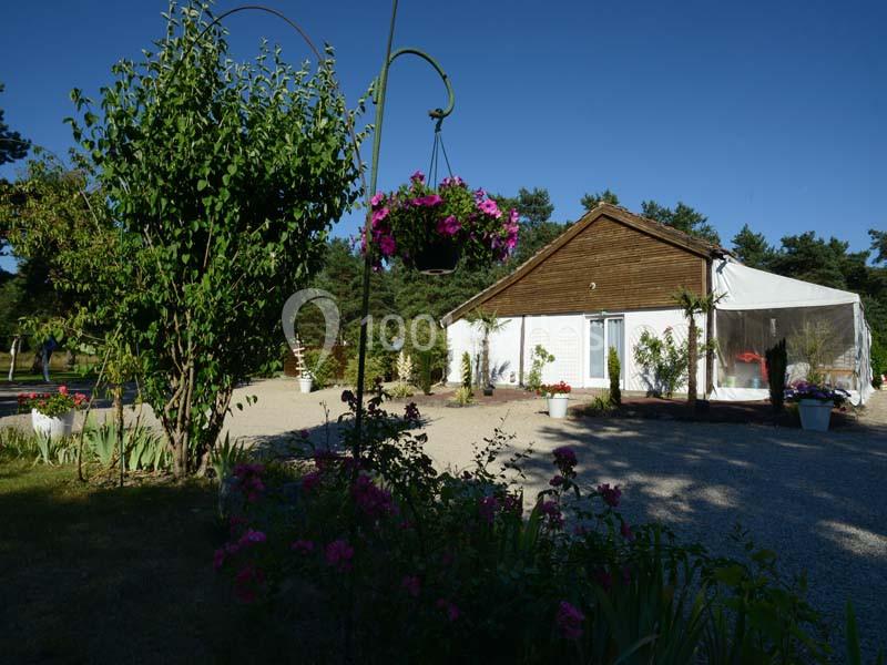 Maison avec façade en bois, entourée de plantes, fleurs suspendues et jardin sous un ciel dégagé.