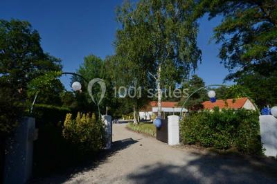 Allée gravillonnée bordée de verdure et de plantes, menant à une entrée avec des arches blanches sous un ciel dégagé.