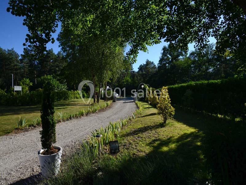 Allée gravillonnée bordée de verdure et de plantes, menant à une entrée avec des arches blanches sous un ciel dégagé.