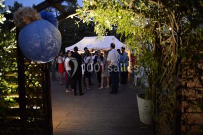 Allée gravillonnée bordée de verdure et de plantes, menant à une entrée avec des arches blanches sous un ciel dégagé.
