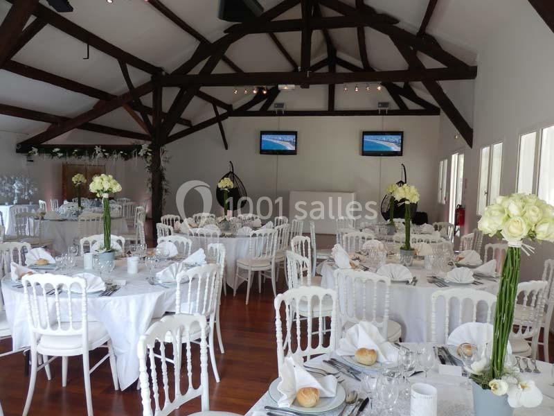Salle de réception lumineuse avec tables rondes dressées, décorée de fleurs blanches et poutres apparentes au plafond.