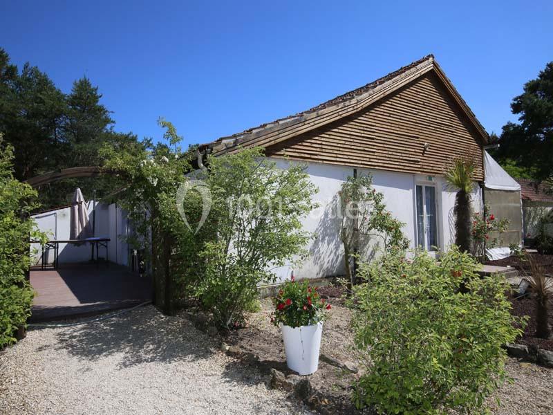 Maison avec façade blanche, toit en tuiles et terrasse ombragée, entourée de végétation et d'un chemin gravillonné.