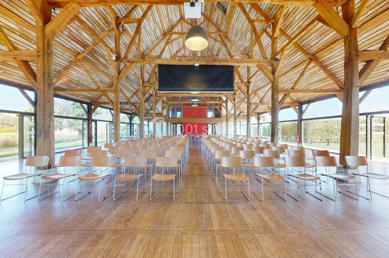 Salle lumineuse avec charpente en bois, rangées de chaises alignées et grandes baies vitrées donnant sur un espace extérieur.