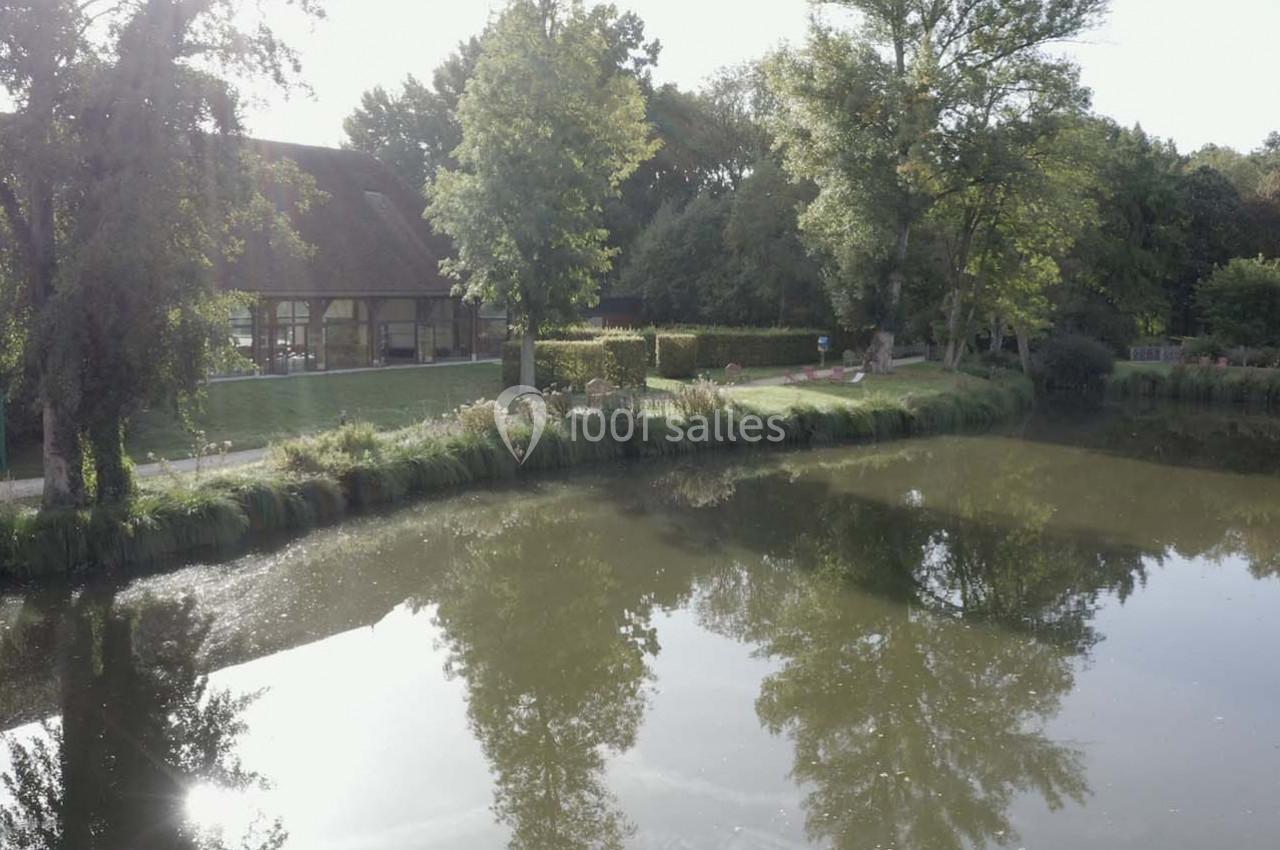 Plan d'eau calme bordé d'arbres et d'une maison à toit en chaume, reflétant le paysage environnant.