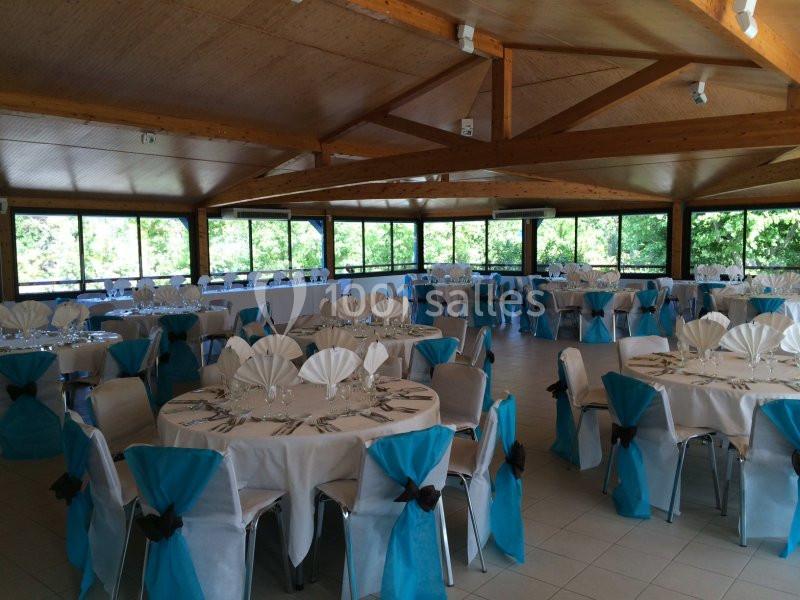 Salle de réception lumineuse avec tables rondes dressées, nappes blanches et chaises décorées de nœuds bleus.