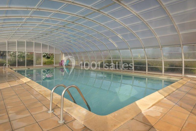 Piscine intérieure couverte par une structure transparente, entourée de carrelage beige et donnant sur un jardin.