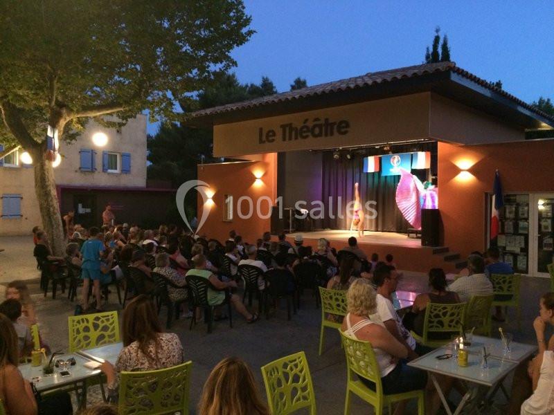 Spectateurs assis en extérieur devant une scène éclairée avec une performance en cours, sous un bâtiment marqué ’Le Théâtre’.
