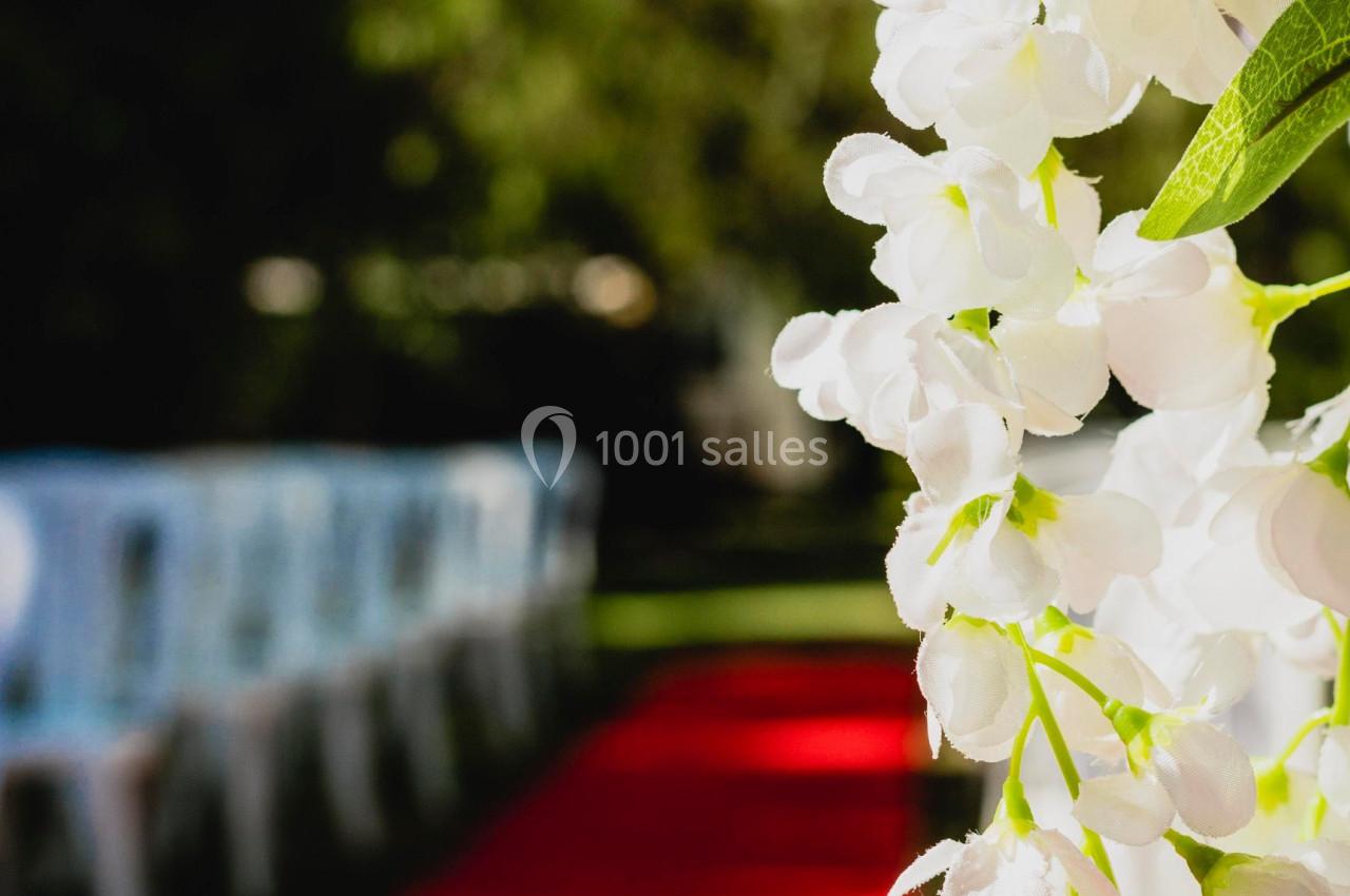 Allée extérieure avec tapis rouge et rangées de chaises blanches, décorée de fleurs blanches au premier plan.