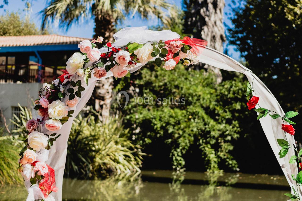 Arche décorée de fleurs roses et blanches devant un étang entouré de végétation et de palmiers.