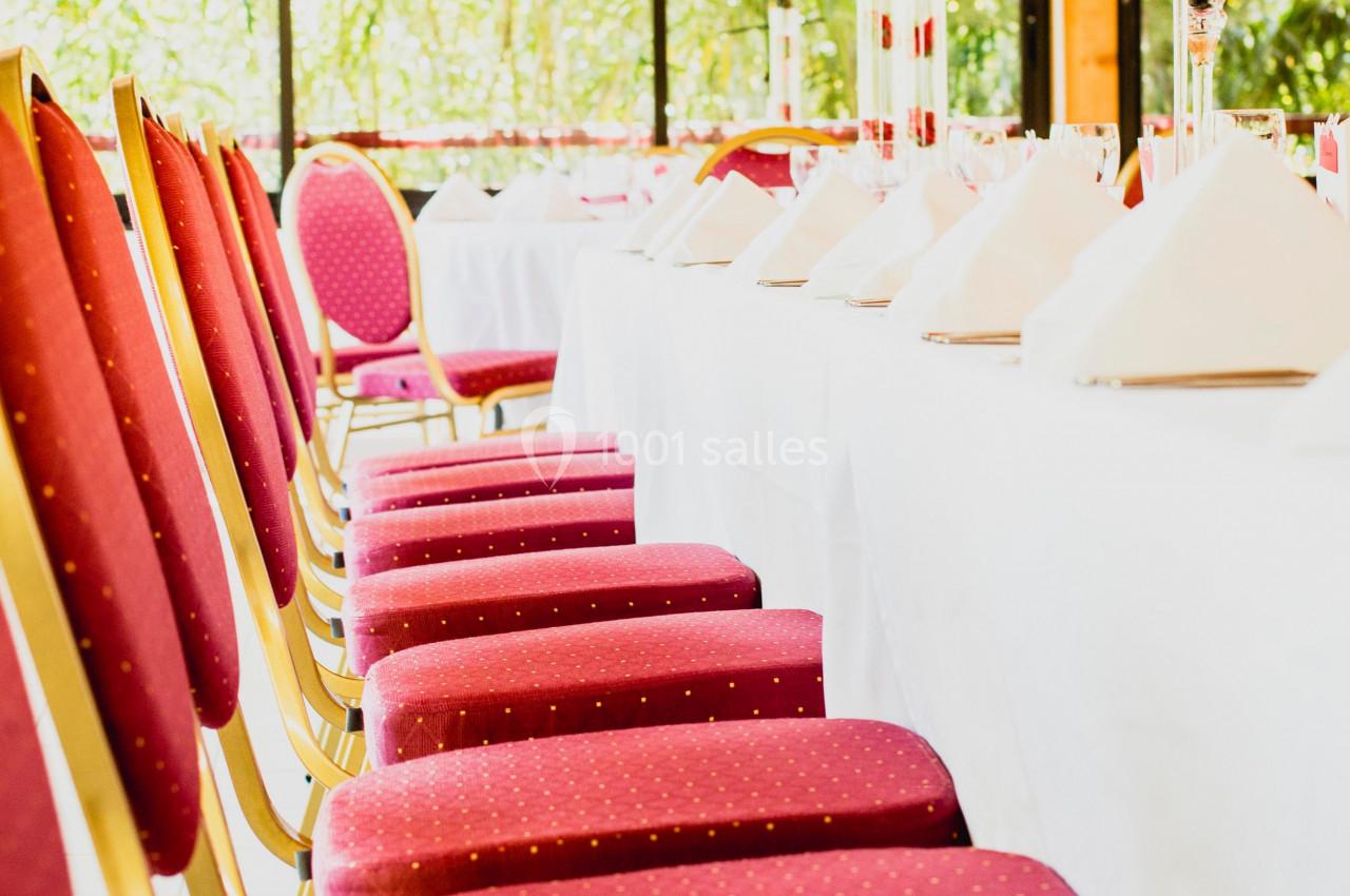 Chaises rouges alignées le long d'une table dressée avec nappes blanches et décorations dans une salle lumineuse.