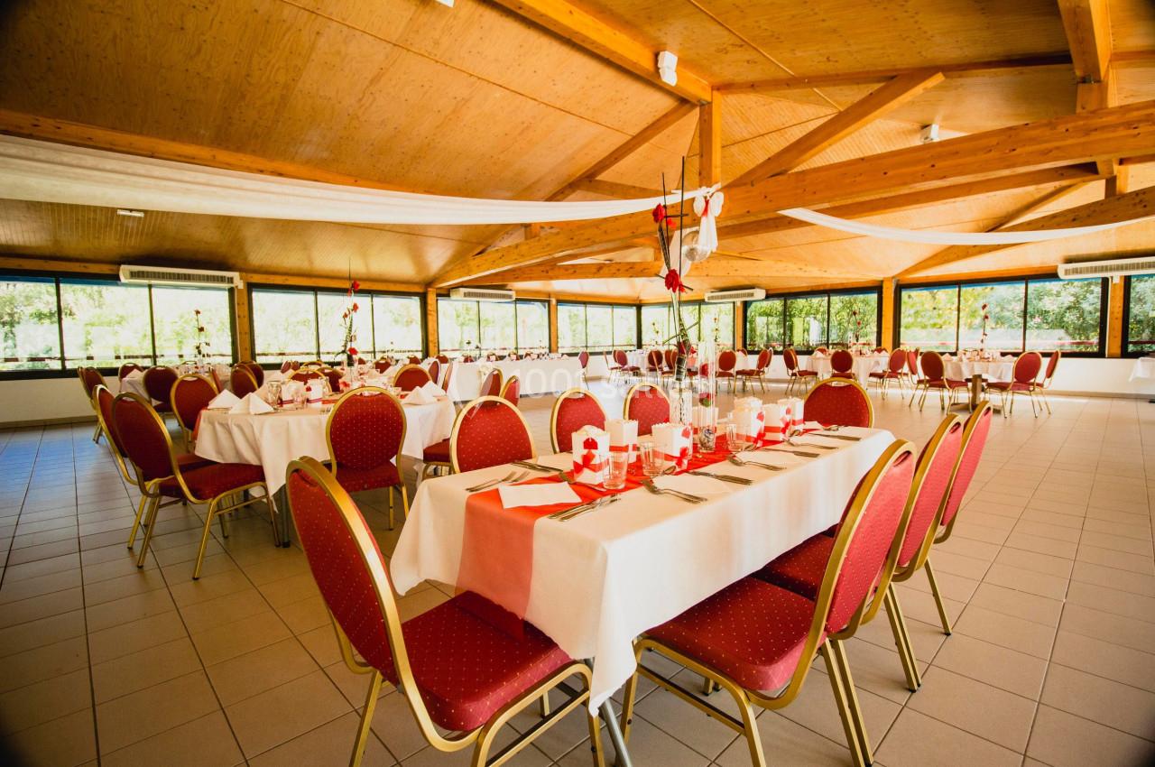 Salle de réception lumineuse avec tables dressées, nappes blanches, chaises rouges et décorations festives.