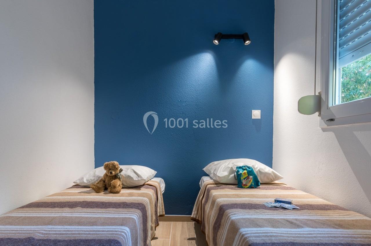 Deux lits simples avec des couvertures rayées, un mur bleu en arrière-plan et une fenêtre partiellement ouverte à droite.