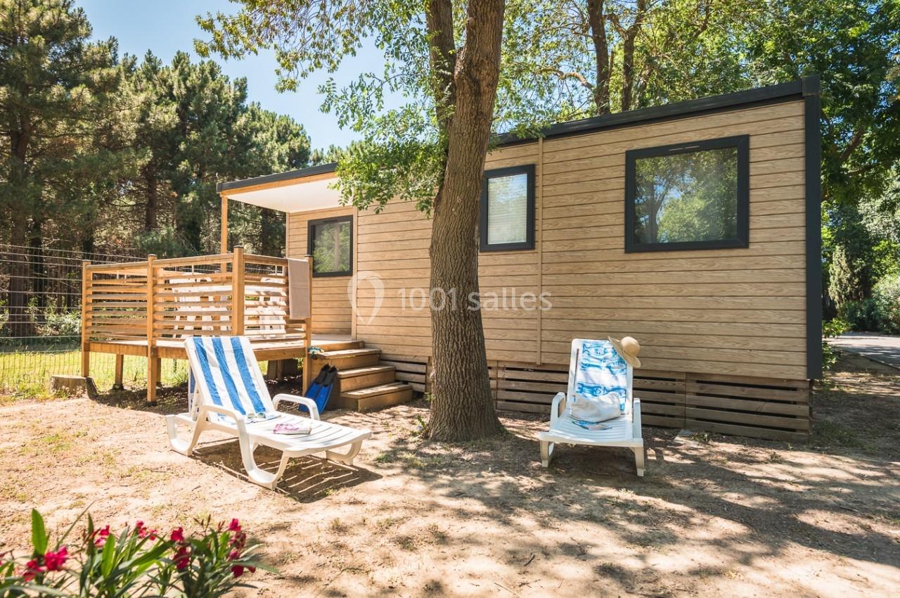 Mobil-home en bois avec terrasse ombragée, entouré d'arbres, deux chaises longues et fleurs au premier plan.