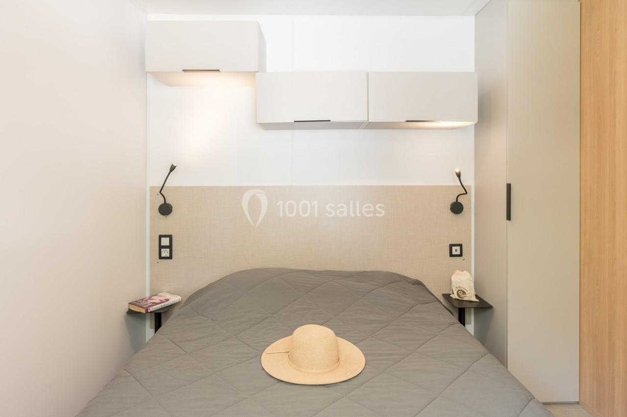 Lit double avec couvre-lit gris, chapeau posé dessus, éclairage mural et rangements suspendus dans une chambre moderne.
