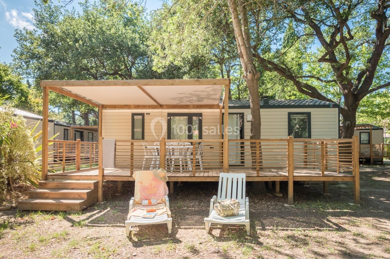 Mobil-home avec terrasse en bois ombragée, entouré d'arbres, équipé de chaises longues et d'une table extérieure.