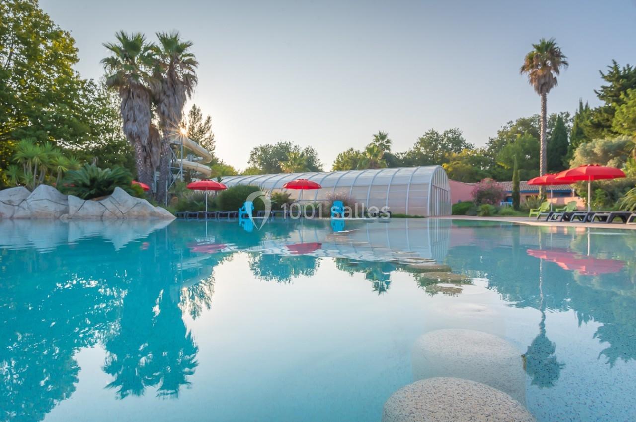 Piscine extérieure avec palmiers, parasols rouges et structure couverte en arrière-plan, sous un ciel dégagé.
