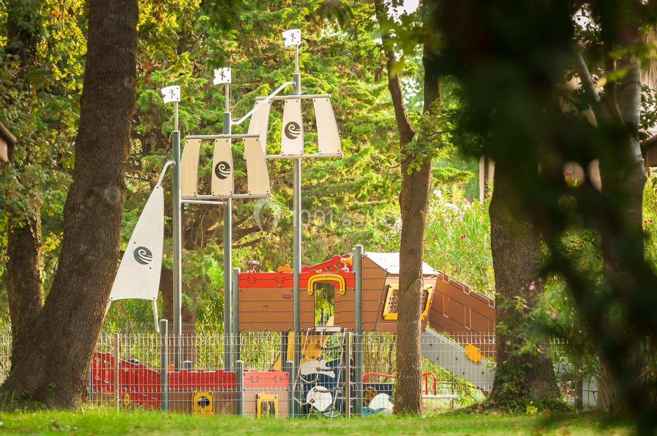 Structure de jeu en forme de bateau pirate entourée d'arbres dans un parc, avec une clôture métallique au premier plan.