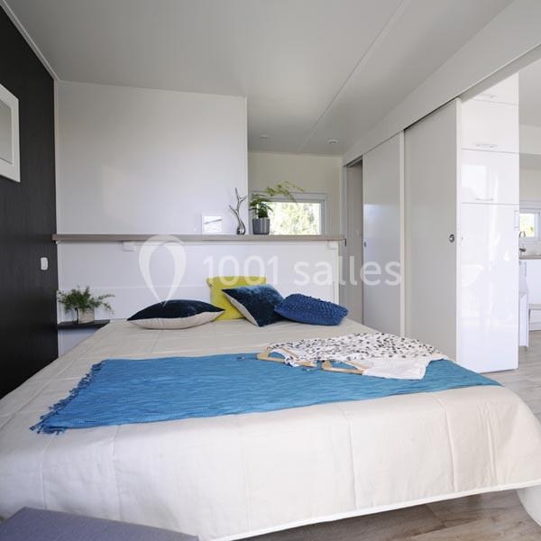 Chambre lumineuse avec lit double, draps blancs, plaid bleu, coussins colorés et placards blancs coulissants.