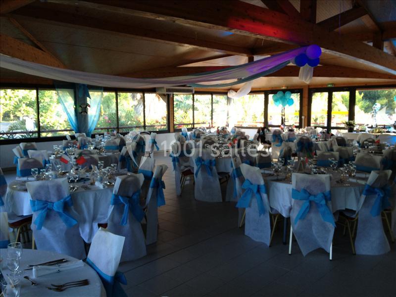 Salle de réception décorée avec des tables rondes, nappes blanches, chaises ornées de rubans bleus et ballons suspendus.