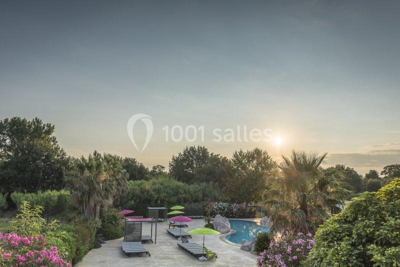 Piscine extérieure entourée de végétation, avec transats et parasols colorés sous un ciel dégagé au coucher du soleil.