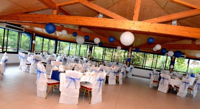 Salle de réception décorée avec des tables rondes, nappes blanches, chaises ornées de rubans bleus et lanternes suspendues.