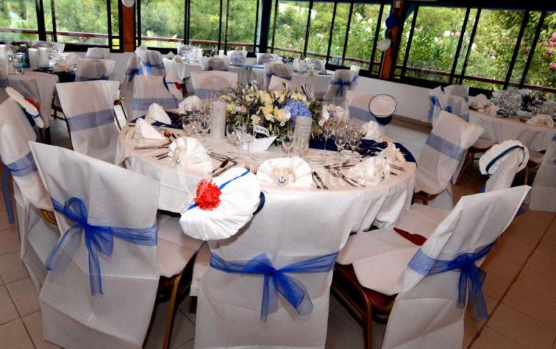 Salle de réception décorée avec des tables rondes, nappes blanches, chaises ornées de rubans bleus et centres de table…