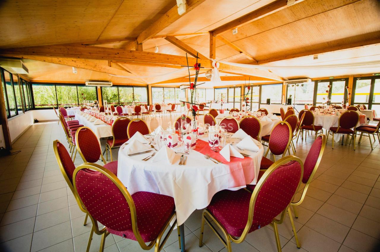 Salle de réception lumineuse avec tables rondes dressées, nappes blanches et chaises rouges, entourée de baies vitrées.