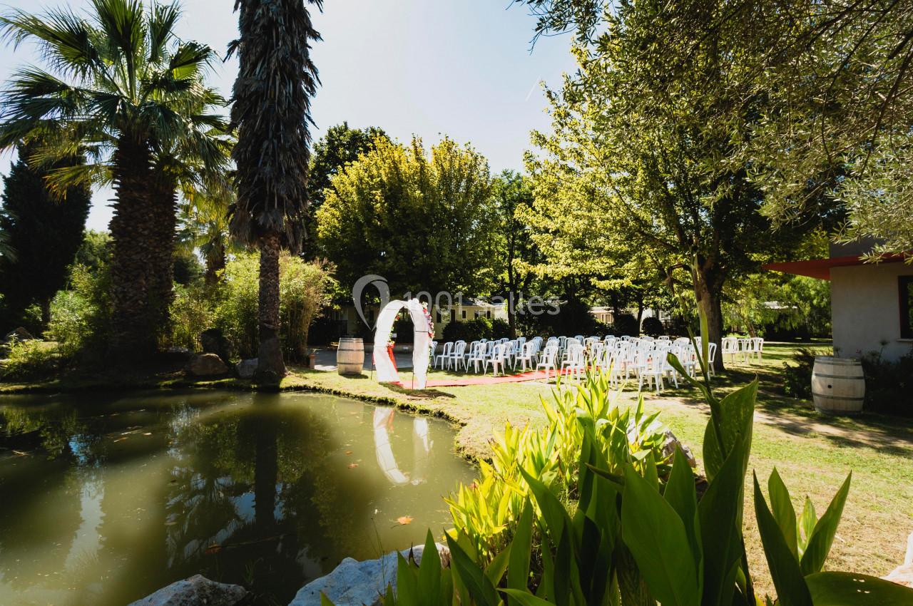 Un jardin avec un étang, des chaises blanches disposées pour une cérémonie et une arche décorative sous les arbres.