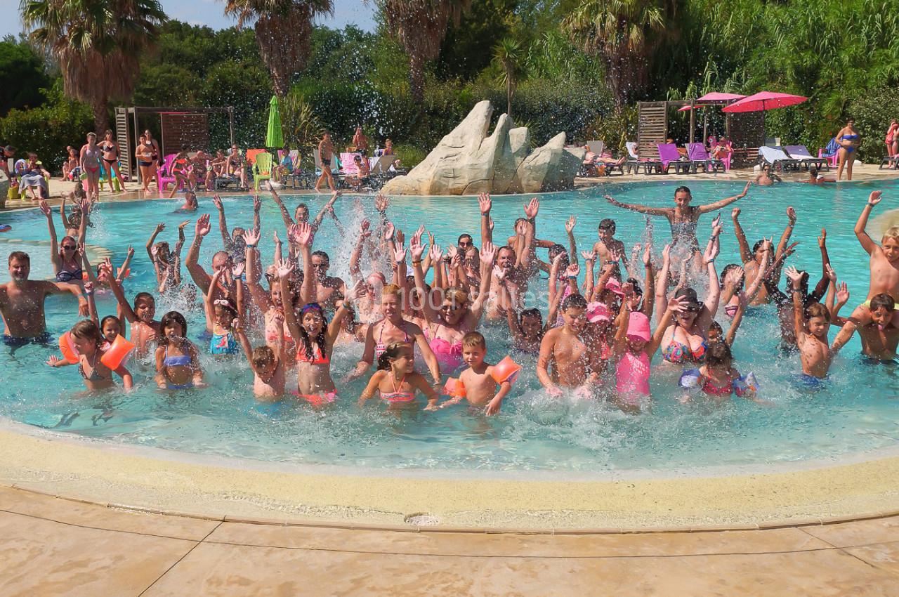 Groupe de personnes s'amusant et éclaboussant dans une piscine extérieure entourée de végétation et de parasols.