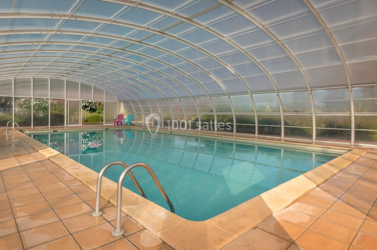 Piscine intérieure couverte par une structure transparente, entourée de carrelage beige et de quelques chaises colorées.