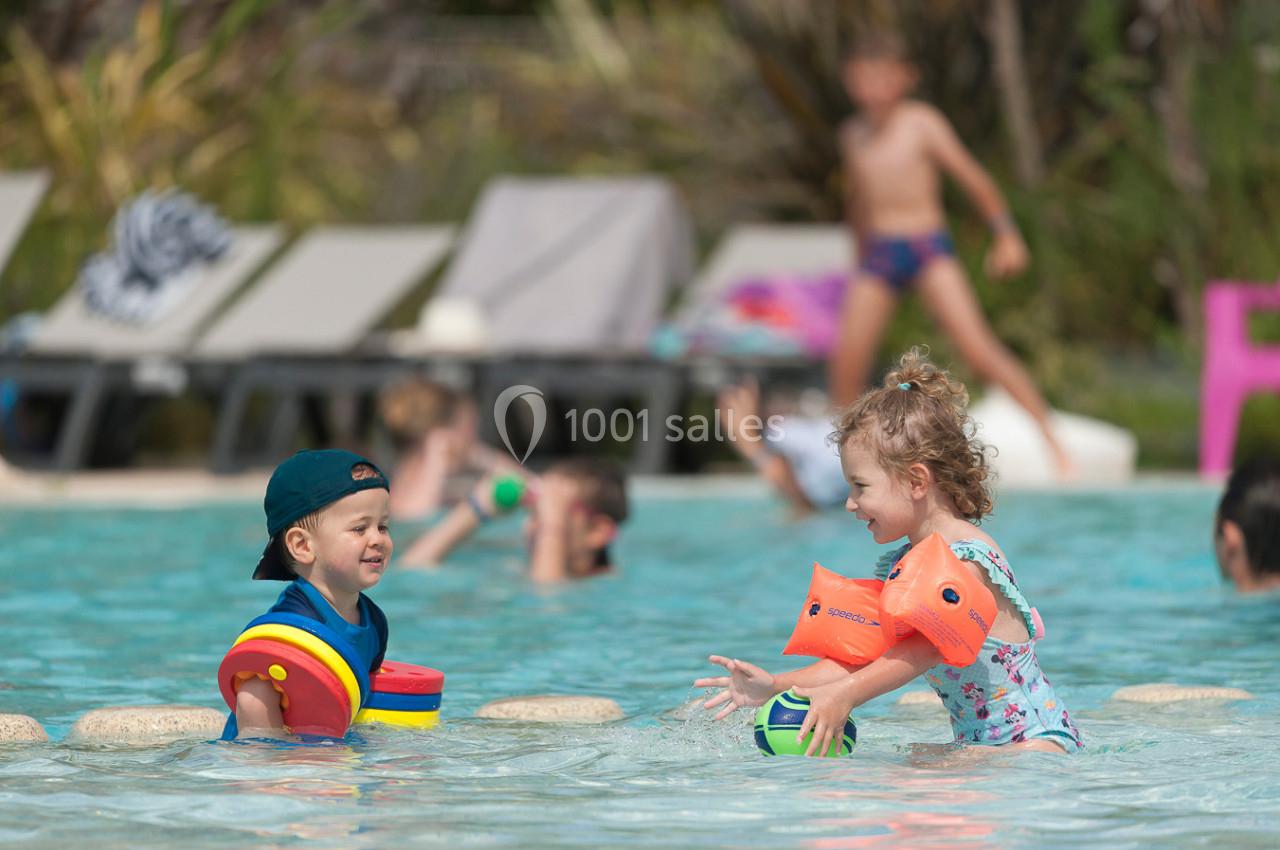 Deux jeunes enfants jouent avec des jouets flottants dans une piscine peu profonde, entourés d'autres baigneurs.