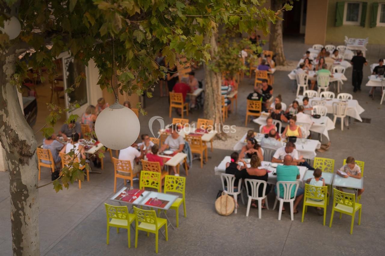 Terrasse extérieure animée avec des tables et chaises colorées, des convives dînant sous des arbres feuillus.