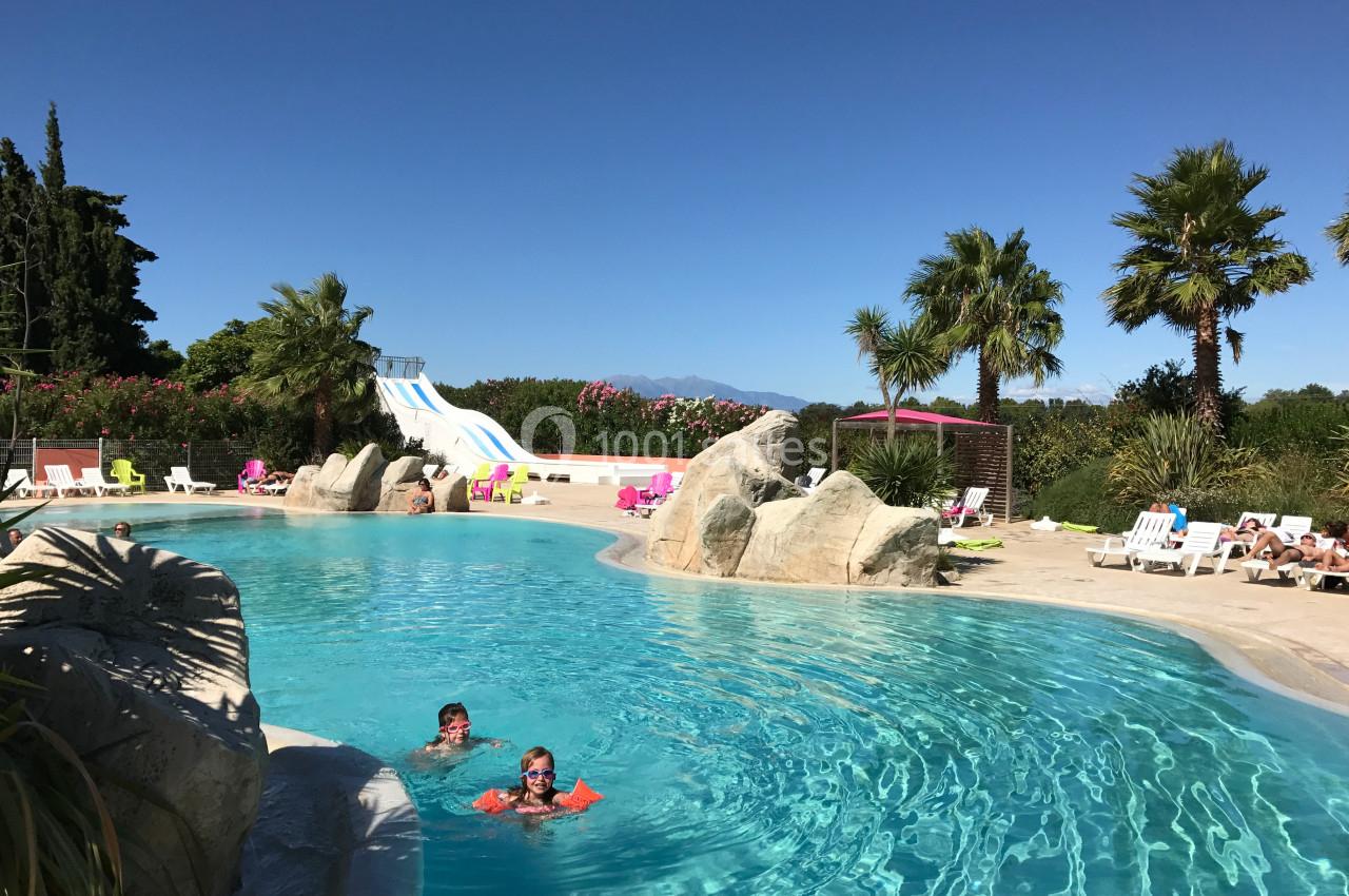 Piscine extérieure avec toboggans, palmiers et transats, deux personnes nageant sous un ciel dégagé.