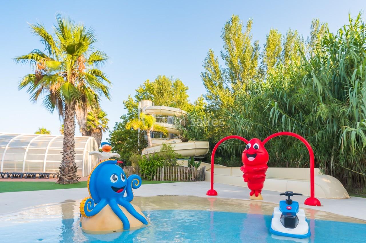 Piscine pour enfants avec jeux aquatiques colorés, toboggan en arrière-plan et végétation environnante.